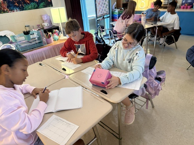Students seated at desks, working on papers and using pens, with one holding a pink box.