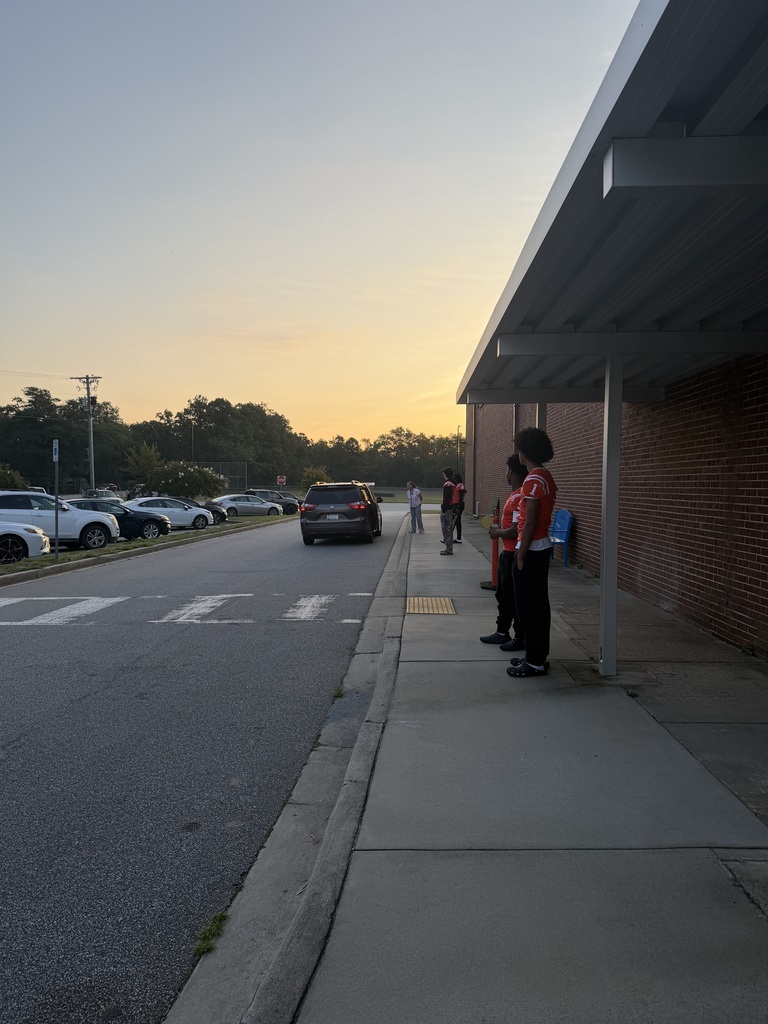 Three people in red shirts and black pants stand on a sidewalk at sunset. A brick building and canopy are on the right. Cars are parked on the street.