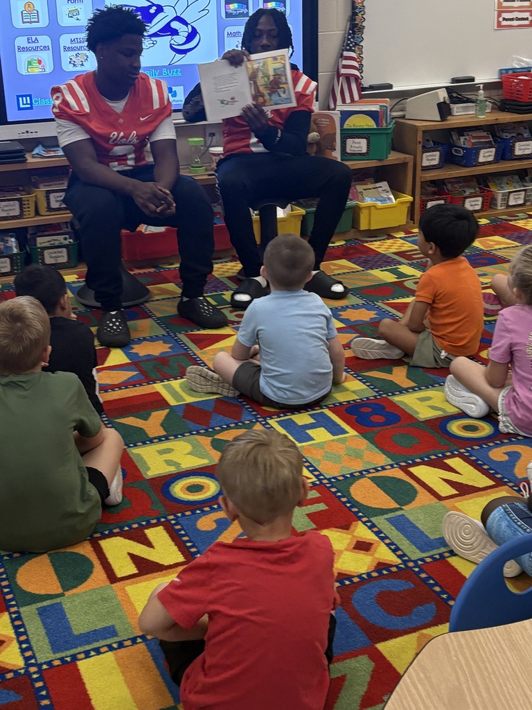 A man reads a book to a group of children sitting on a carpeted floor. Behind them is a classroom with a monitor.