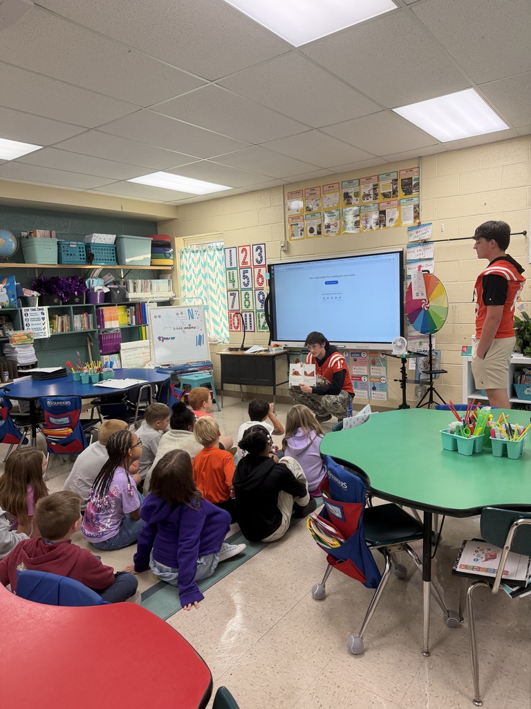 A classroom with a teacher reading to students. A large screen is on the wall, and shelves with books are in the background.