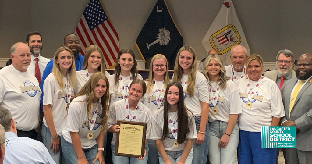 A group stands in front of flags. They wear white T-shirts, some with medals. A person holds a framed certificate.