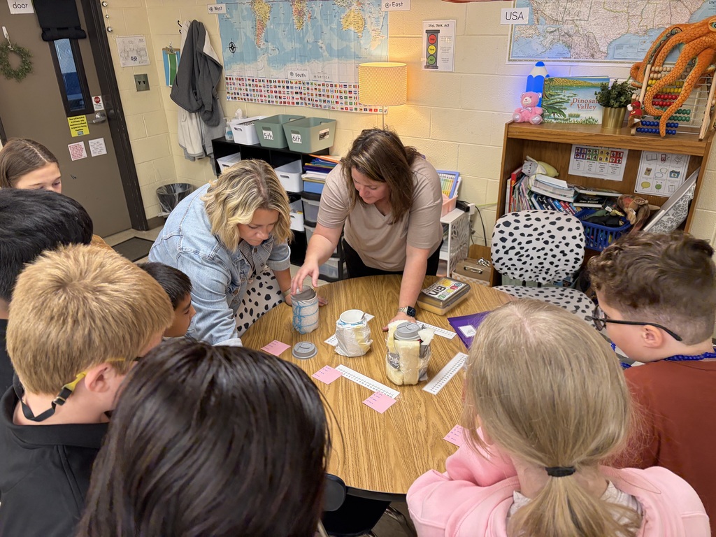 Several children sit around a table with two adults, who are showing them a science experiment.