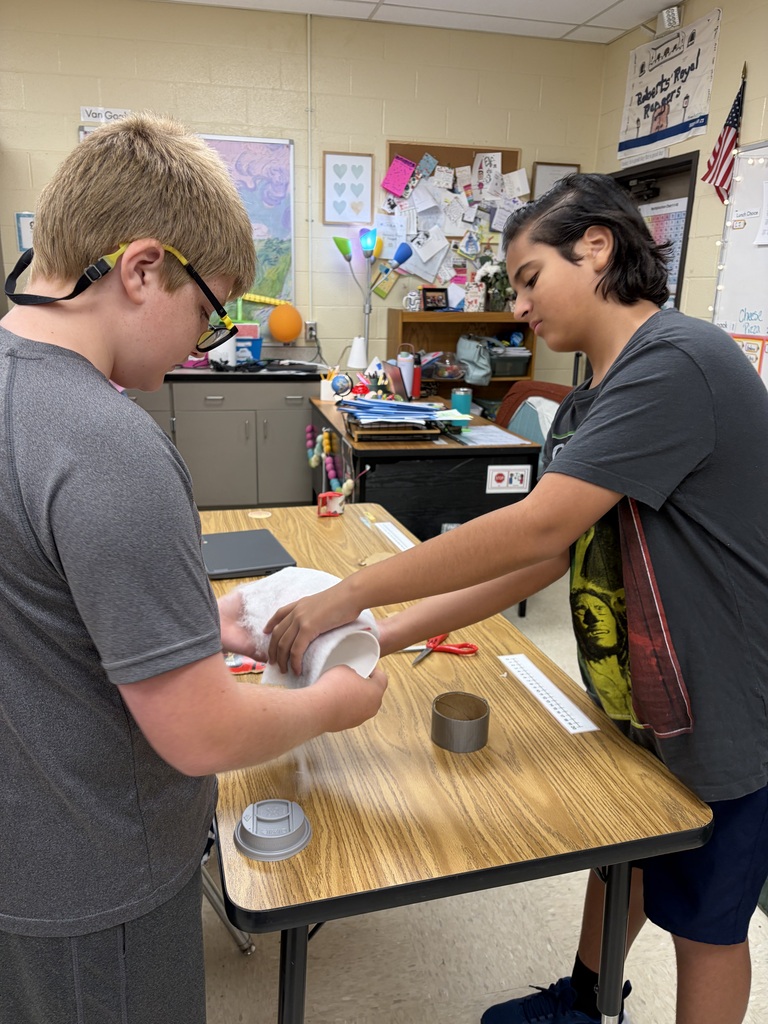 Two students in a classroom work on a project at a table, handling materials and tools.