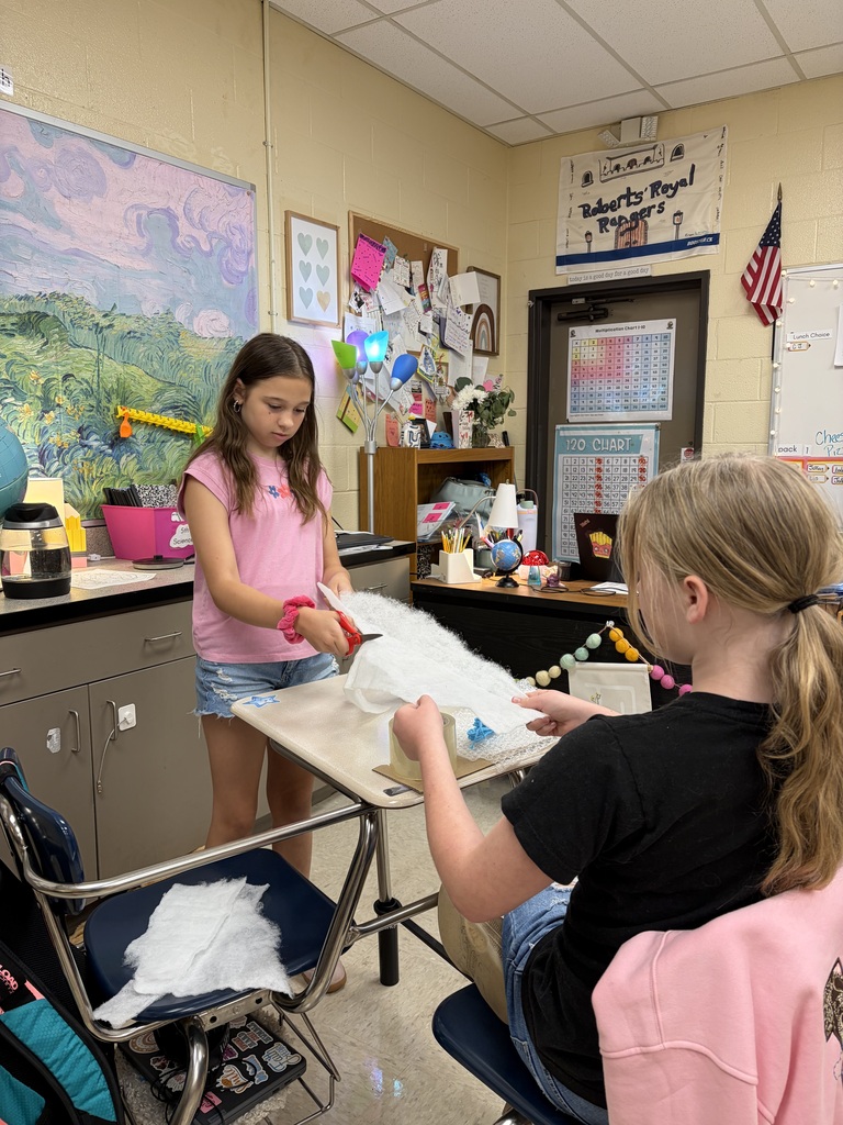 Two girls seated in a classroom, one standing at a desk cutting fabric, the other sitting and working on the cut fabric.
