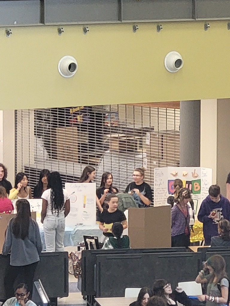 Many individuals at an event, possibly an exhibition, with displays and tables; some wear glasses and bracelets.