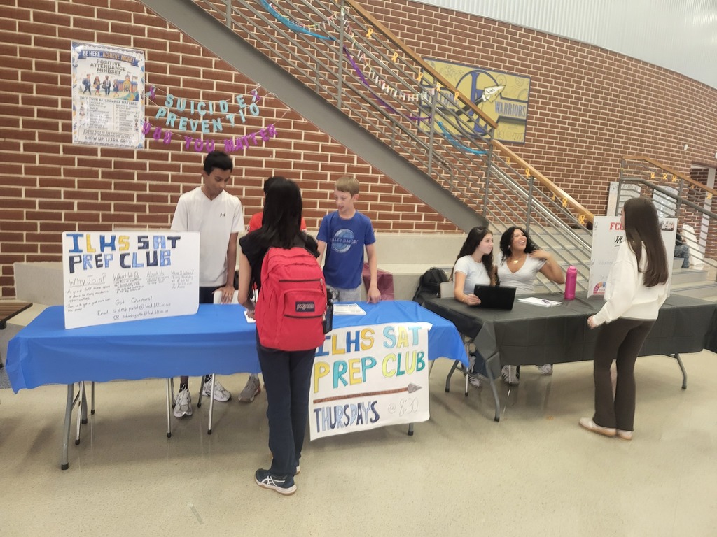 Indoor area with tables and people. Tables have banners with "IHS SAT PREP CLUB". People stand near tables, one with a backpack. A brick wall and stairs in the background.