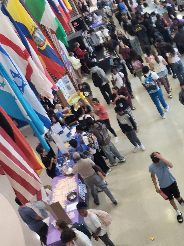Many people, some with bags, walk around a hall with flags from different countries hanging.