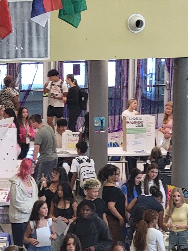 A crowded room with a yellow wall. Many people are standing and talking. A camera is mounted on the wall.