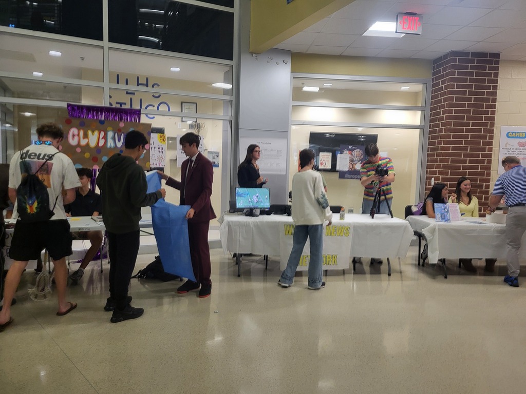 Indoors, people gather around tables. A monitor and a camera are on one table. Two pillars are in the background.