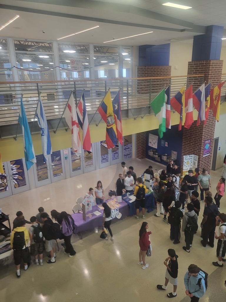 Many people gather in a school hallway. Tables with purple tablecloths are set up. Flags hang from the ceiling.