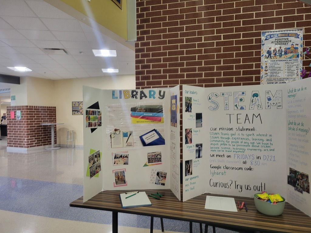 A display in a hallway with a sign reading "Library STEAM Team" on a wooden table. The backdrop is a brick wall.