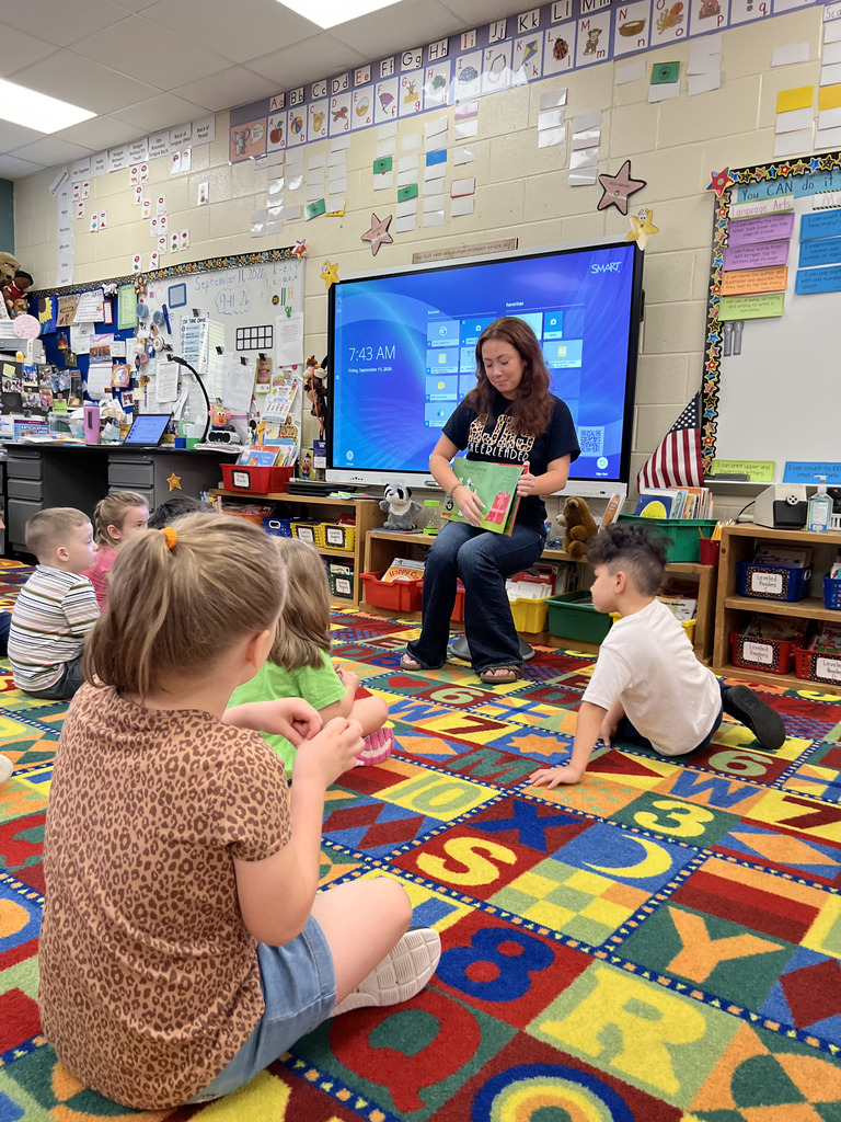 A woman holds a book while sitting in front of a classroom screen. Children sit on a colorful floor mat.