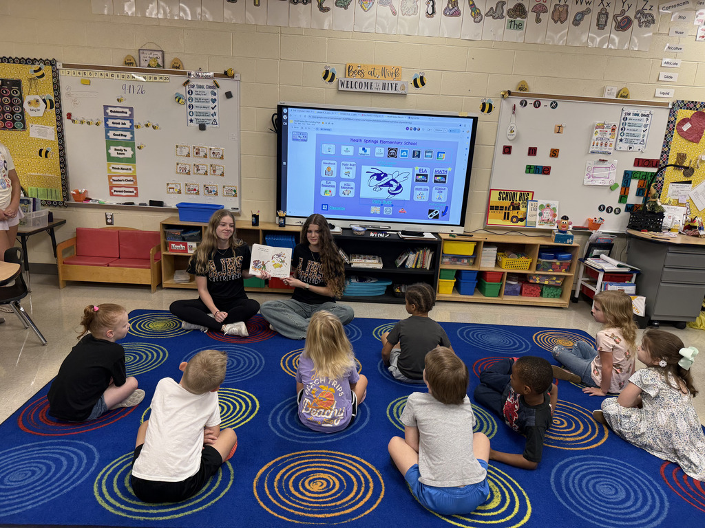 A group of young children seated in a circle around two teachers, listening to a story.