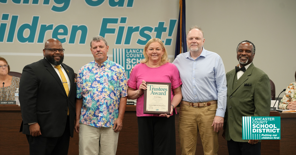 Five adults stand with an award plaque. The woman in the center holds it. A banner behind them reads "Children First!"