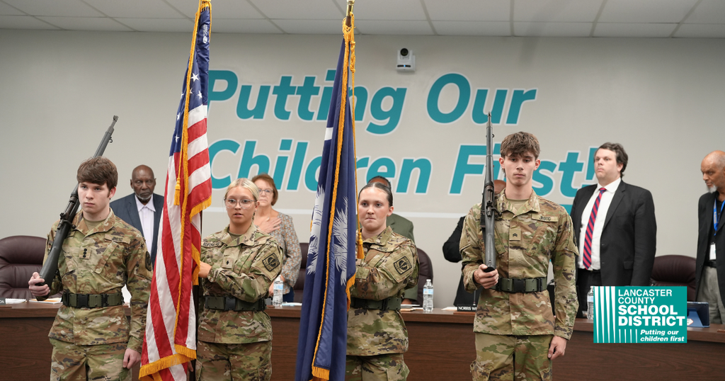 Four people in uniform hold flags in front of a table with a slogan. A meeting is in progress.
