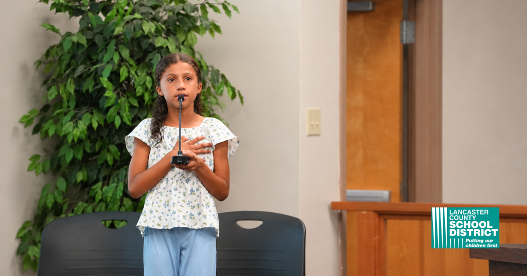 A girl stands and speaks into a microphone, with a plant and wooden door behind her.