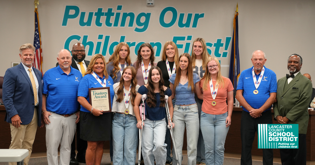 A group of students and adults stand for a photo. All wear medals. Behind them is a white wall with text.