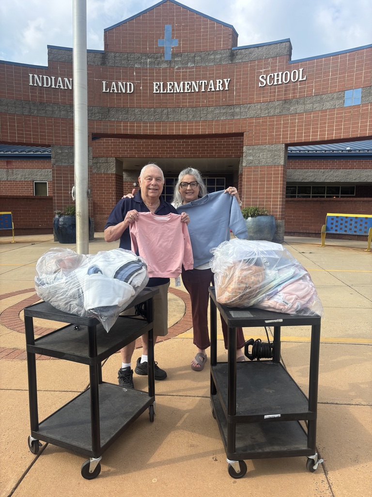 Two people stand next to carts holding packages in front of a brick building. A sign above reads "Indian Land Elementary School."