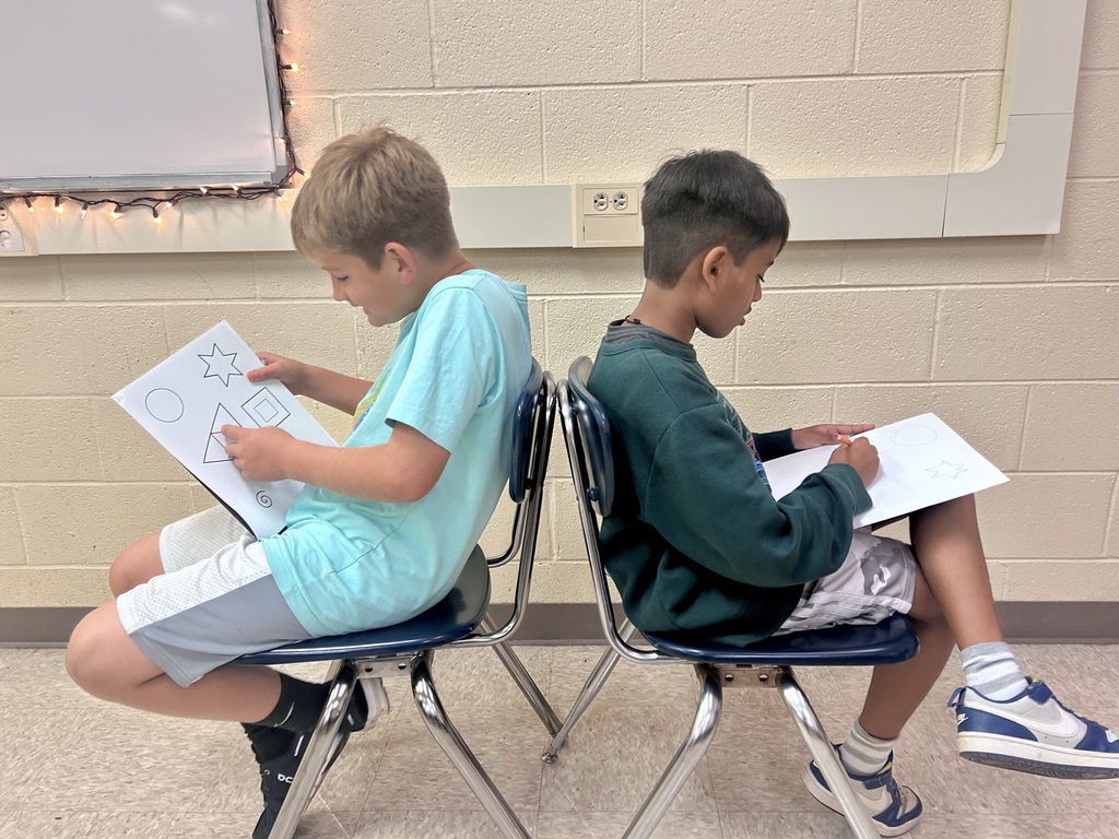 Two young boys sit in chairs and look at papers in front of them. One holds a pen.