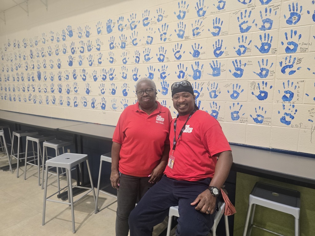 Two men in red shirts and hats pose in front of a wall with blue handprints.