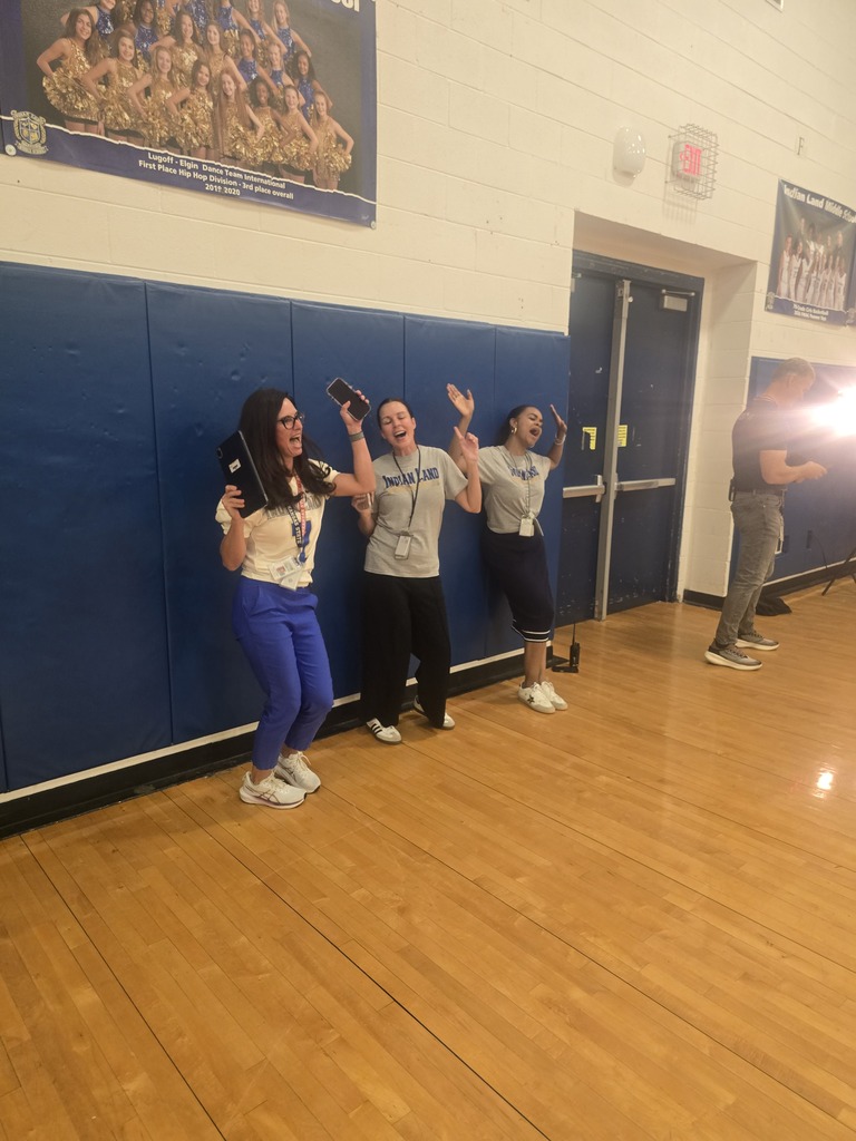 Three people are standing on a wooden floor in a gym. One person is raising a hand.
