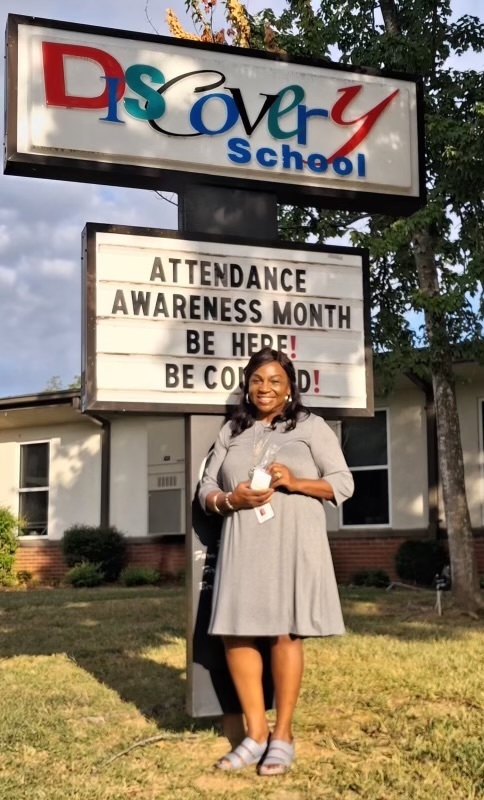 Attendance clerk standing in front of school sign holding a gift cards.