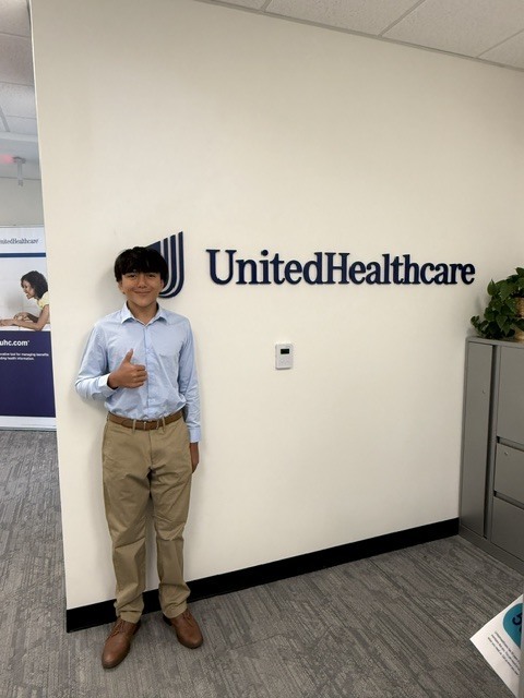 A man in a blue shirt and khaki pants stands in an office, giving a thumbs up.