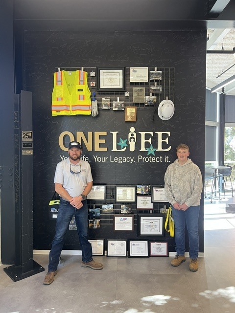 Two people stand in front of a wall with a sign and various safety equipment.