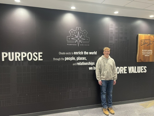 A person in a white hoodie and blue jeans stands in front of a black wall with white text.