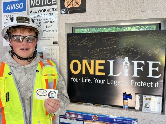 Person wearing safety vest, helmet, and glasses holds a small card in front of a sign.