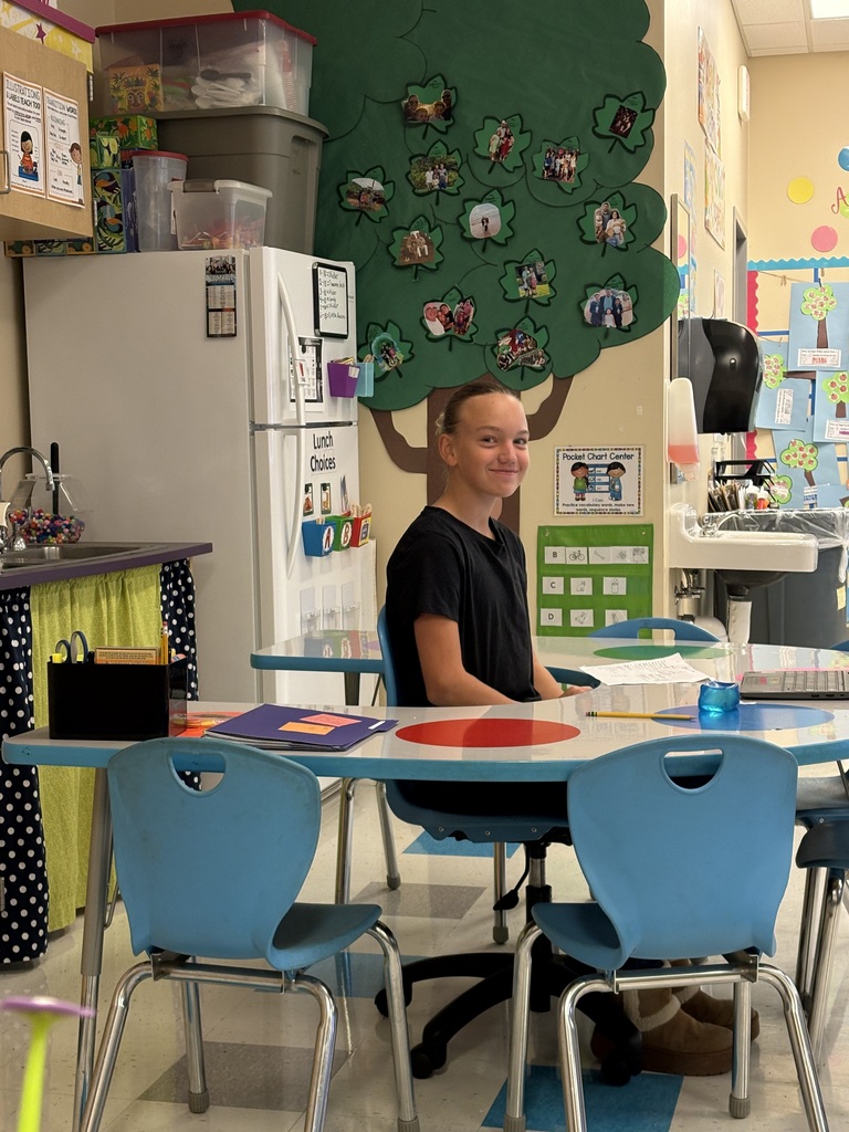 A student sits at a desk with books and papers, near a refrigerator and a tree mural.