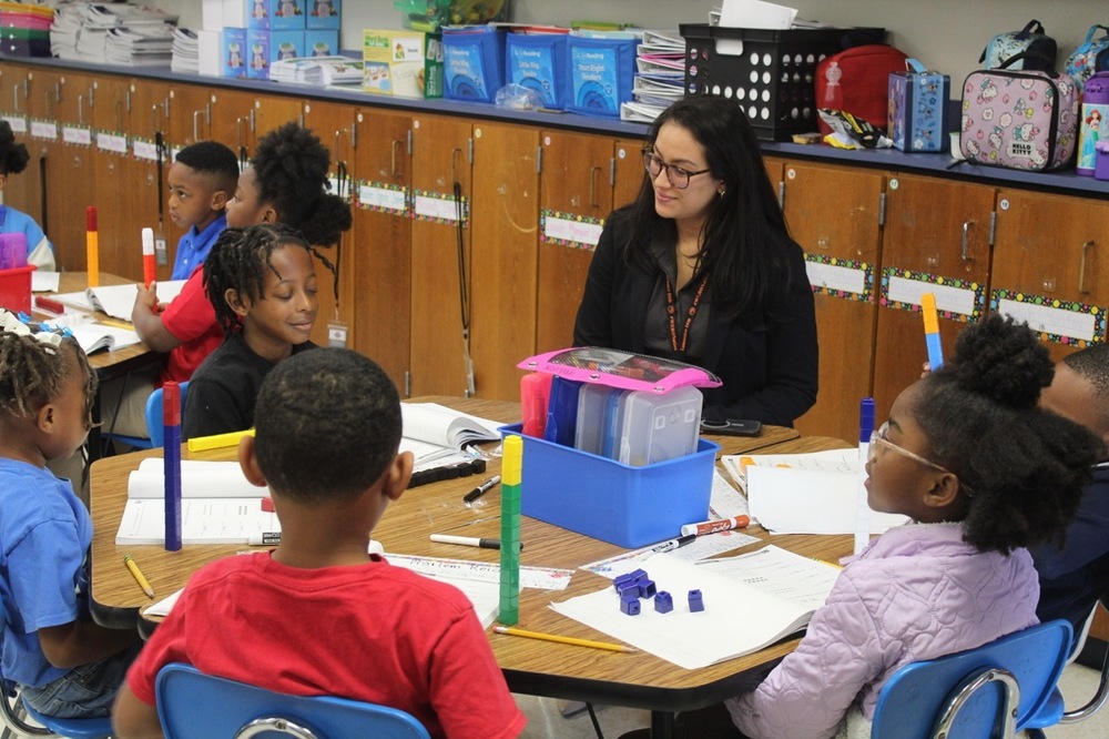 Adult woman sitting at table with five students during math class