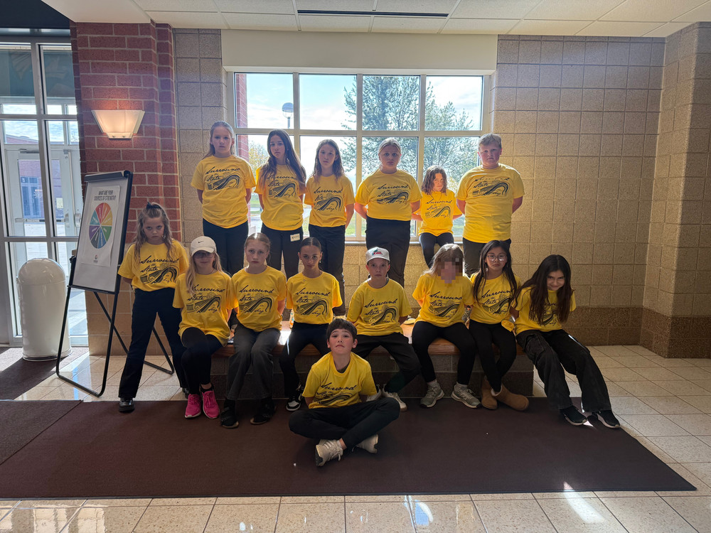 A group of fifteen students wearing matching bright yellow T-shirts with a black graphic and text that reads “Langdon Area” and features a design of a running figure. They are seated and standing in two rows inside a building with beige tiled walls and large windows in the background. A sign with a colorful circular chart is displayed on an easel to the left.
