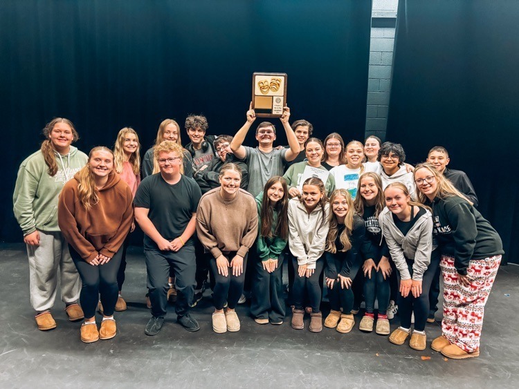 A large group of people standing on a stage with a black backdrop. One person in the center is holding a wooden plaque with two gold theatrical masks raised above their head. The group is arranged in rows, with some standing and others leaning forward.
