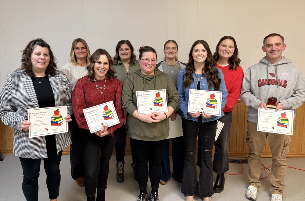Alt text: A group of nine individuals standing indoors against a light-colored wall with a wooden lower panel. Eight people are holding certificates featuring a colorful stack of books with an apple on top, and one person on the far right is holding both a certificate and a small award. The group is arranged in two rows, with five in the front and four in the back. The floor is light gray, and an orange extension cord is visible near the wall on the right side.