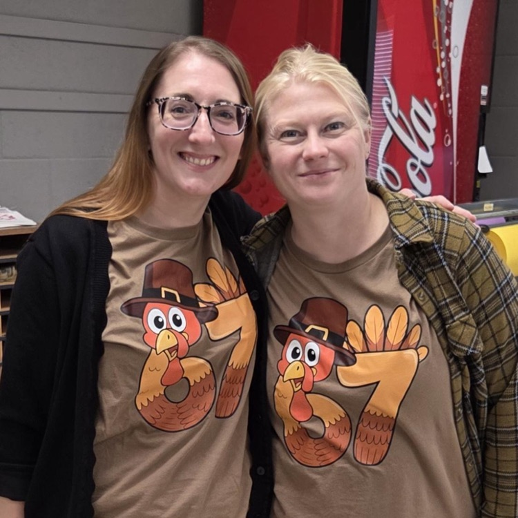 wo individuals wearing brown shirts with a Thanksgiving-themed design. The shirts feature a cartoon turkey wearing a pilgrim hat, with the number “67” integrated into the turkey’s body. They are standing near a red vending machine.