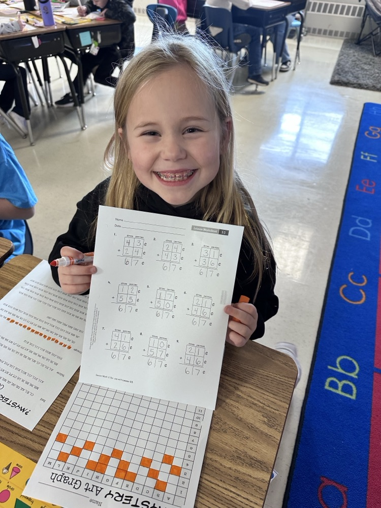A student seated at a desk holding up a worksheet with math problems involving fractions (6/7). The desk has additional papers and a colorful graphing activity. The classroom floor has a rug with alphabet letters.