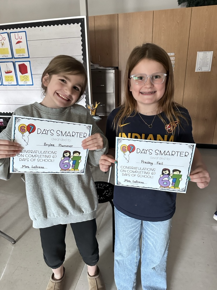 Two students standing in a classroom holding certificates that read “67 Days Smarter” with colorful graphics of numbers and cartoon characters. The certificates congratulate them for completing 67 days of school. Behind them is a bulletin board with alphabet cards.