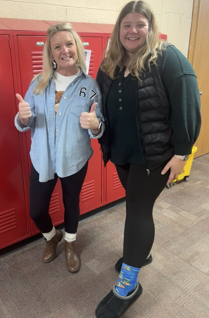 Two individuals standing in a hallway in front of red lockers. One is wearing a light blue button-up shirt over a white top with the number “67” printed on the chest, black leggings, and brown ankle boots, giving a thumbs-up gesture. The other is wearing a dark green top, a black puffer vest, black leggings, and black slip-on shoes with bright blue patterned socks. A yellow cleaning cart is visible in the background near a wooden door.