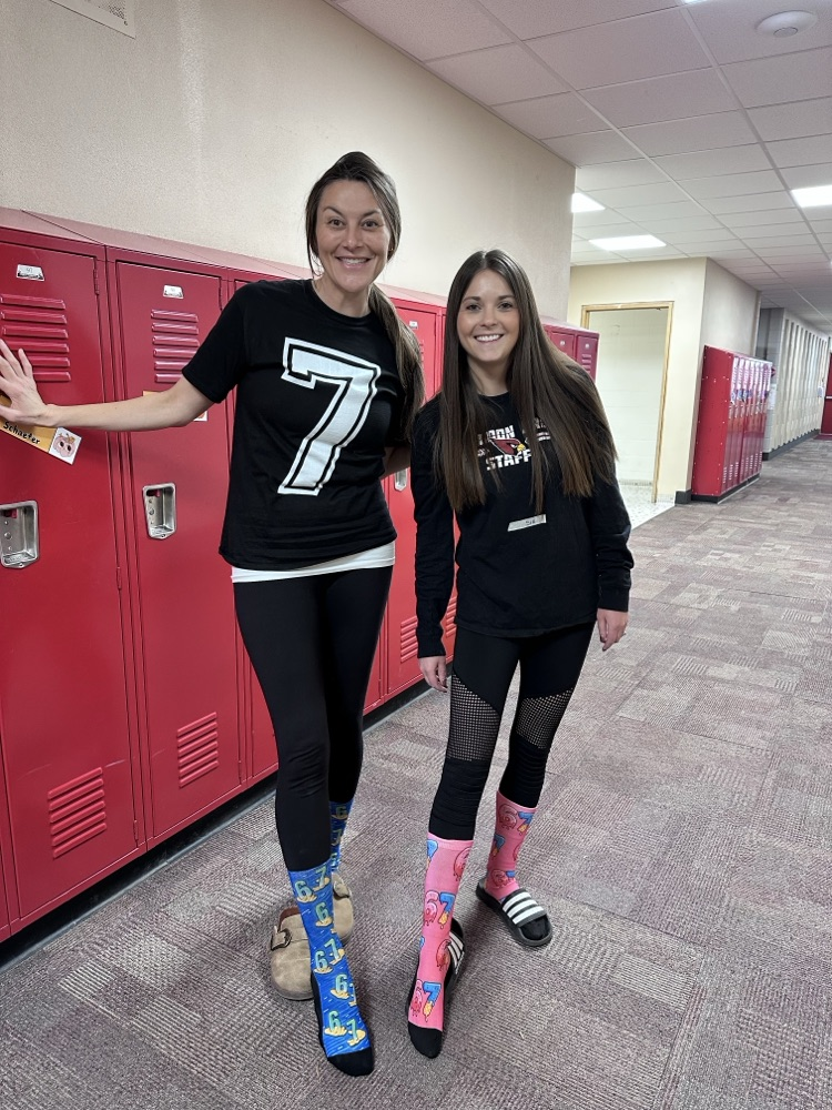 Two individuals standing in a hallway with red lockers. One is wearing a black shirt with a large white number “7,” and the other is wearing a black shirt with text and graphics. Both have colorful mismatched socks and casual footwear.