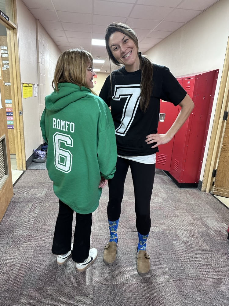 Two individuals in a hallway with red lockers. One is wearing a black shirt with a large white number “7,” and the other is wearing a green hoodie with the name “ROMFO” and the number “6” printed on the back. Both are wearing casual pants and shoes.
