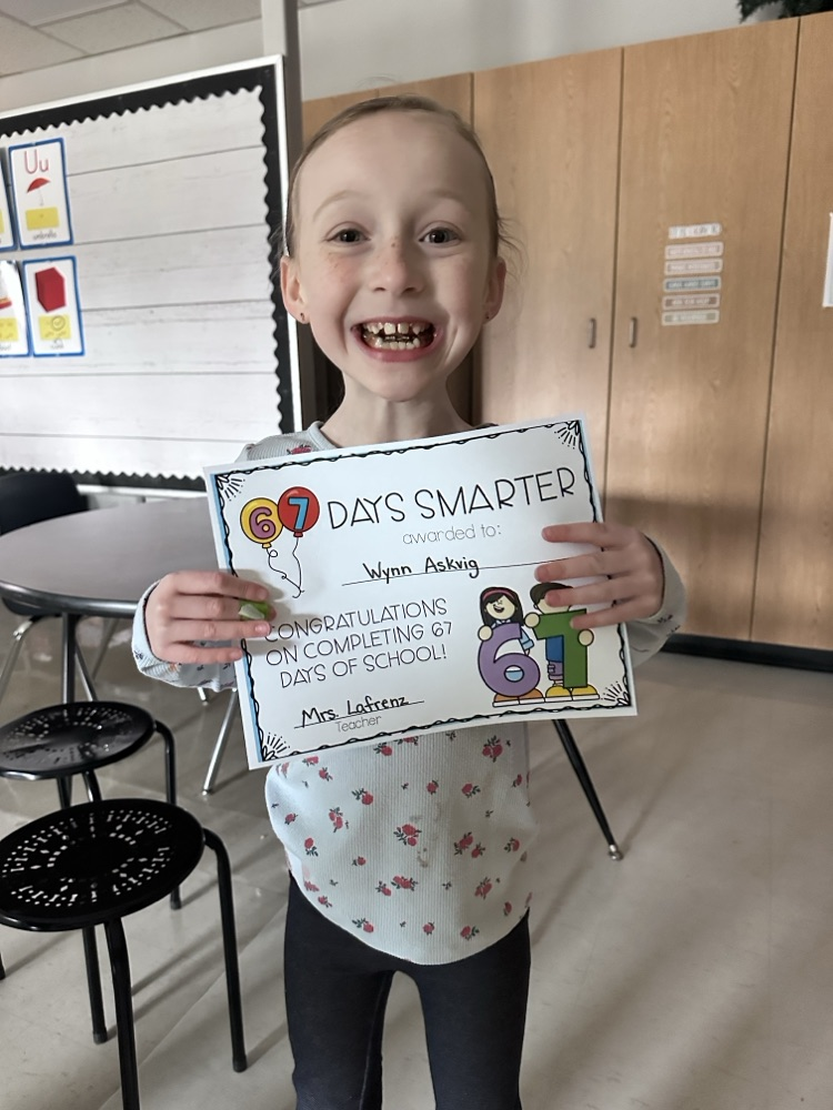 A student holding a certificate that reads “67 Days Smarter” with colorful graphics. The certificate congratulates the student for completing 67 days of school. The classroom background includes tables, chairs, and a bulletin board.