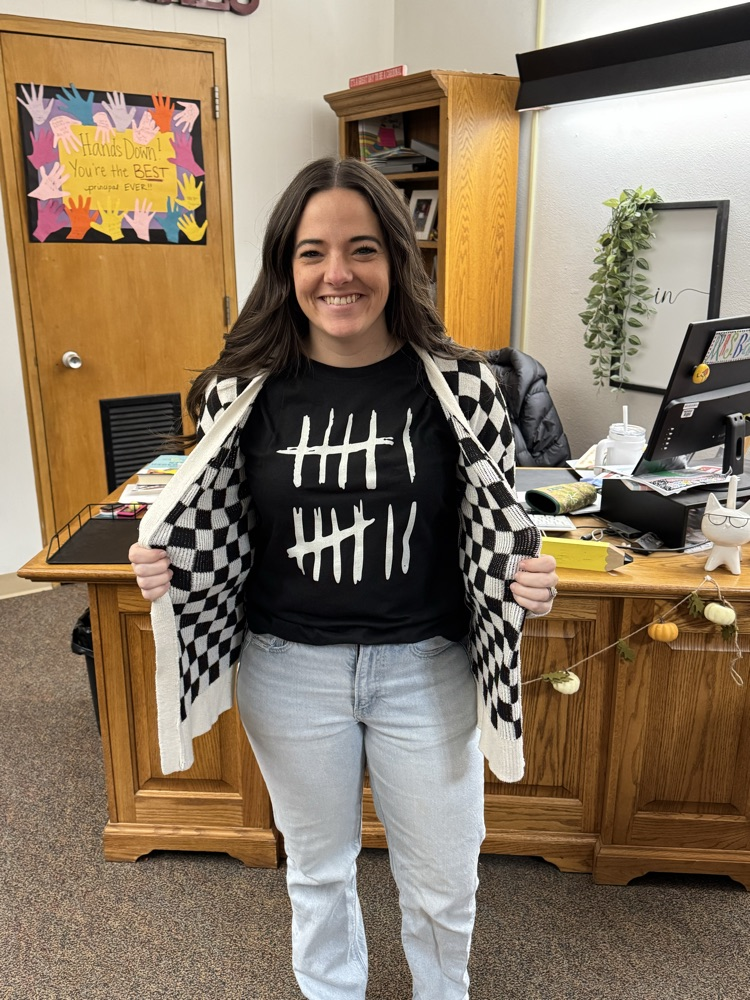 An individual standing in an office wearing a black shirt with tally marks representing the number 67. The person is also wearing a black-and-white checkered cardigan and jeans. Behind them is a wooden desk with office supplies and a door decorated with colorful handprints.