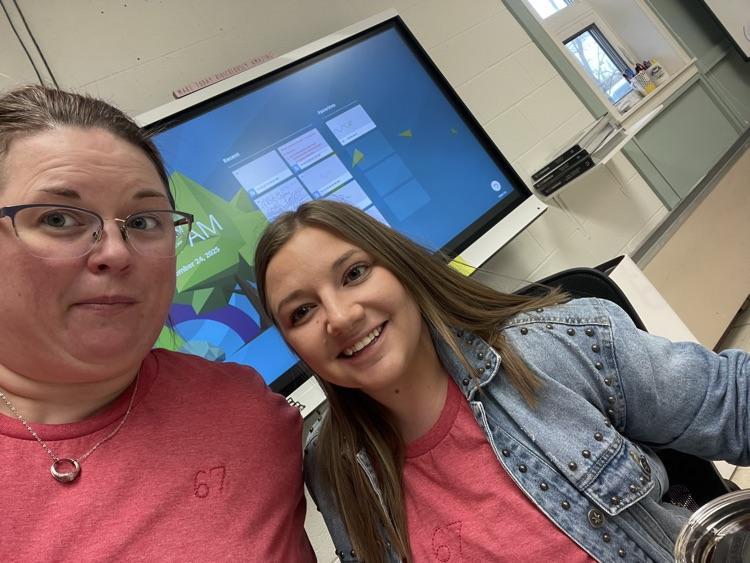 Two individuals wearing matching red shirts with the number “67” stitched on the front. They are seated in a classroom with a large interactive display screen in the background showing a colorful interface. The room has white walls, a window, and shelves with supplies.