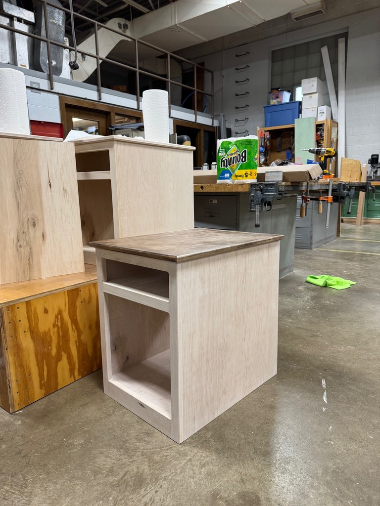 Unfinished wooden nightstand made of light-colored maple sitting on a concrete floor in a workshop. The nightstand has an open storage space and a smaller compartment above it. Behind the nightstand are other wooden pieces and workshop items, including paper towels, a box of Bounty paper towels, clamps, and tools on a workbench.