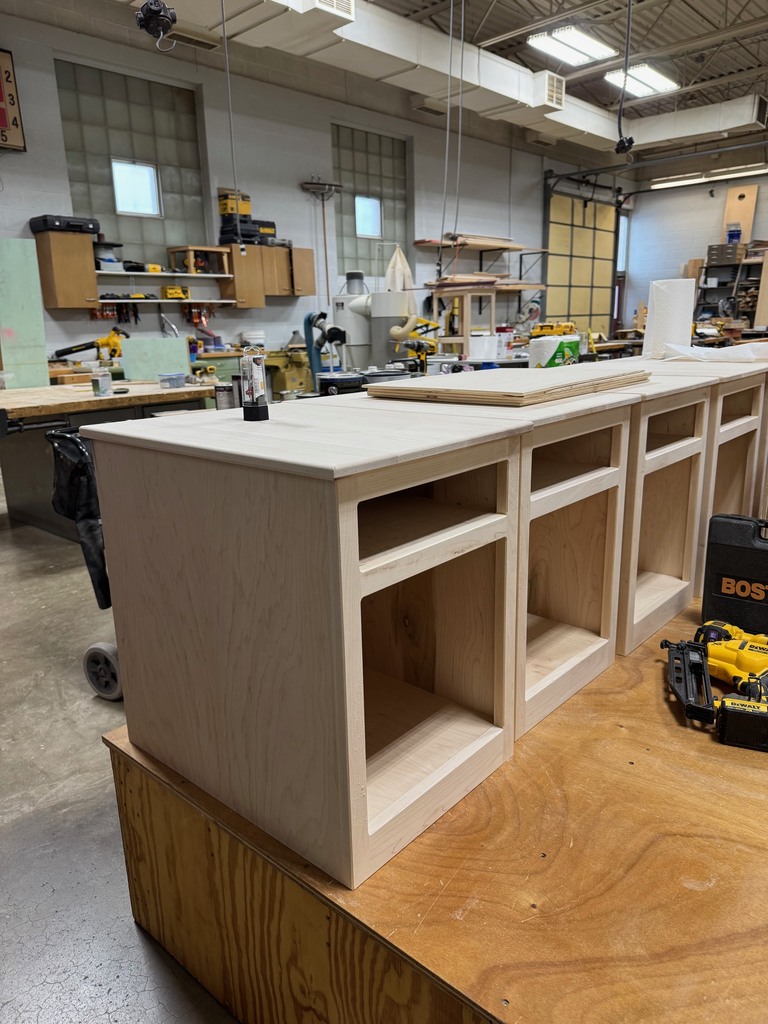 Unfinished wooden nightstand made of light-colored maple resting on a workbench in a workshop. The nightstand has an open storage space and a smaller compartment above it. Several similar nightstands are lined up behind it. In the background, there are worktables, tools, clamps, and woodworking equipment scattered throughout the shop.