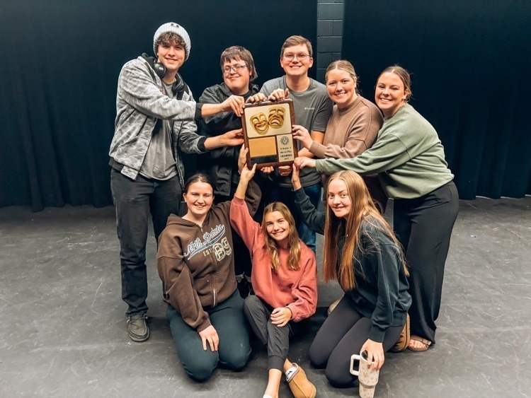 A group of eight people standing and kneeling on a stage with a black backdrop, holding a wooden plaque featuring two gold theatrical masks. The group is gathered closely, with several hands touching the plaque.