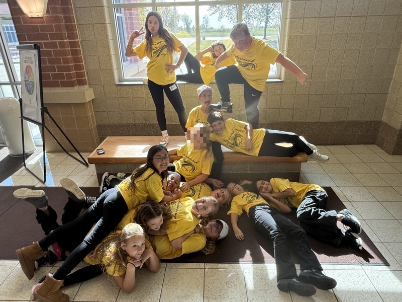 A group of students wearing matching bright yellow T-shirts with a black graphic and text, gathered in a playful pose inside a building with beige tiled walls and large windows. Some students are lying on the floor in a pile, while others are sitting on a bench or standing on and around it. A sign on an easel with a colorful circular chart is visible to the left, and sunlight streams through the window in the background.