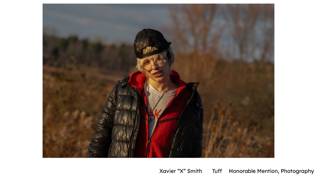 Xavier “X” Smith – “Tuff” (Photography) Alt text: A portrait photograph of a person standing outdoors in a field during golden-hour light. The subject wears a black puffer jacket, red hoodie, and backward cap, with dry grasses and trees softly blurred in the background.
