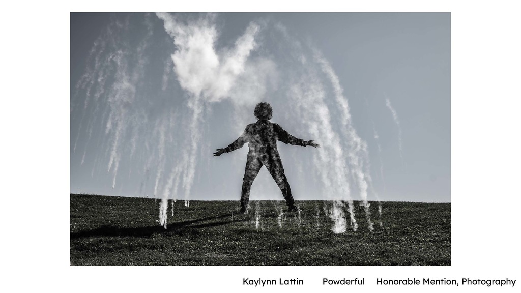 Kaylynn Lattin – “Powderful” (Photography) Alt text: A black-and-white photograph showing a silhouetted person standing on a grassy hill with arms outstretched. White powder bursts into the air around the figure, creating dramatic cloud-like shapes against a bright sky.